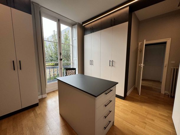 Dressing élégant avec armoires blanches et noires sur toute la hauteur, îlot central avec plateau noir, parquet en bois clair, éclairage LED intégré et une grande fenêtre laissant entrer la lumière naturelle.